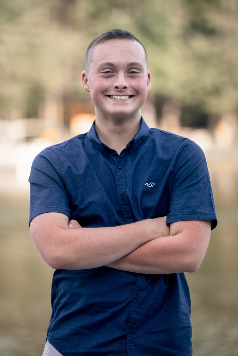 Young man in blue button-up shirt with arms crossed, smiling at camera with blurred outdoor background